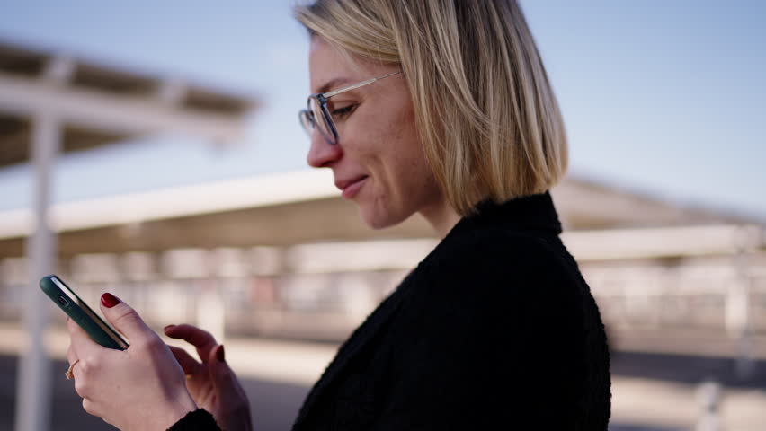 Contemplative young Caucasian business woman focused on her smartphone, reviewing important work emails or finalizing travel plans, standing on airport parking lot with soft daylight | Shutterstock HD Video #1111792137
