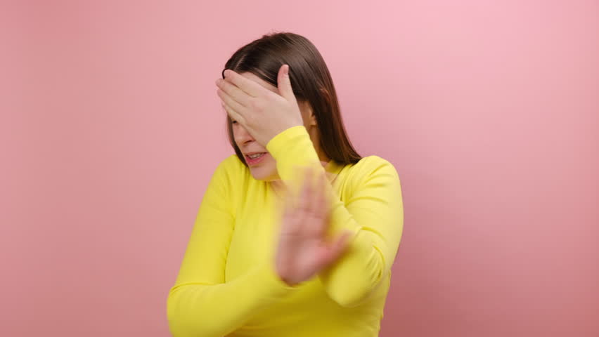 Portrait of young brunette woman keeping hand on eyes, grimacing in disgust and turning away from camera, avoiding watching something shameful. posing isolated on pink color background wall in studio | Shutterstock HD Video #1111792321
