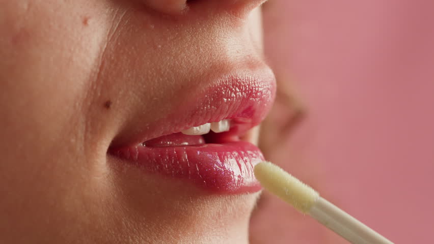 Beauty lip care. Young woman applying hygienic lipstick on lips close-up on pink background. Make-up routine