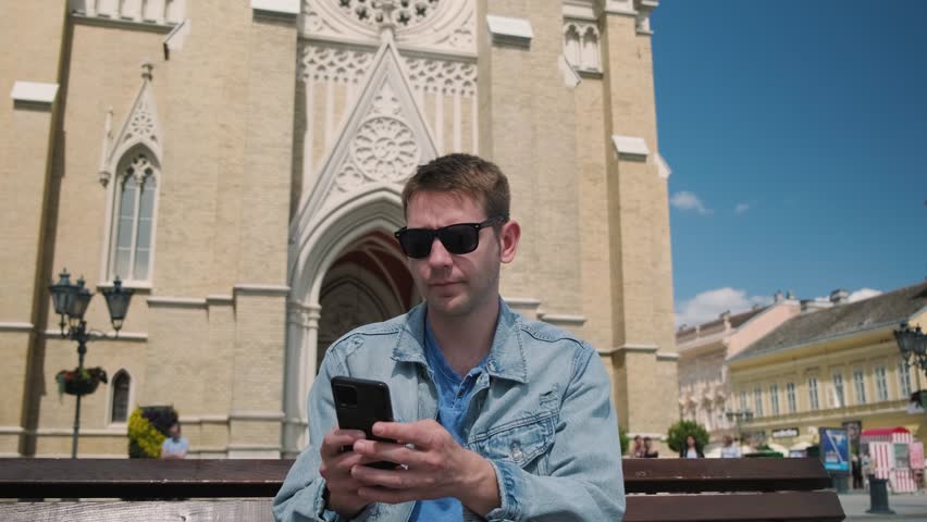 Man tourist sits on a bench in the city square with mobile phone and booking hotel from online application on his phone. Male traveler looking for last minute booking. Travel concept.