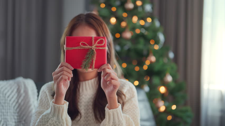 Happy attractive asian woman holding and showing a red gift to the camera. smile woman sitting near a decorated tree with Christmas decorations, Christmas Eve, New Year