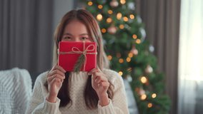 Happy attractive asian woman holding and showing a red gift to the camera. smile woman sitting near a decorated tree with Christmas decorations, Christmas Eve, New Year - Powered by Shutterstock - Get 15% off with code: PIKWIZARD15