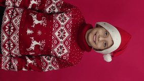 happy Caucasian schoolboy 9 years old in a sweater and Santa hat triumphantly raises his hands up, looks at the camera. studio shooting, red background. vertical video
