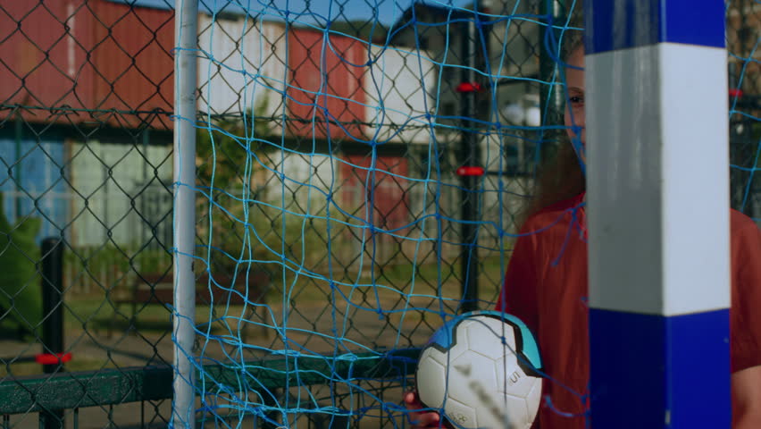 Young girl in sports uniform pulls back net with hand to better see players coming to court. Teenage girl stands on football field with soccer ball in grasp and gazes into distance closeup