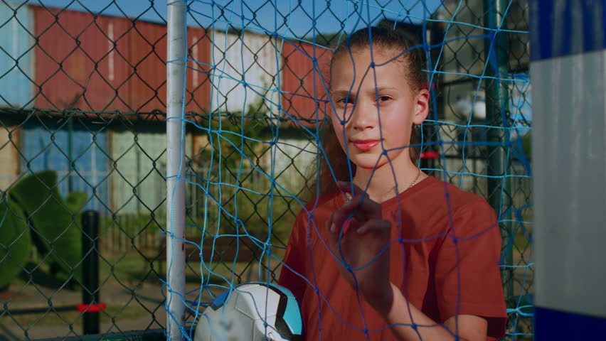 Young girl in sports uniform pulls back net with hand to better see players coming to court. Teenage girl stands on football field with soccer ball in grasp and gazes into distance closeup