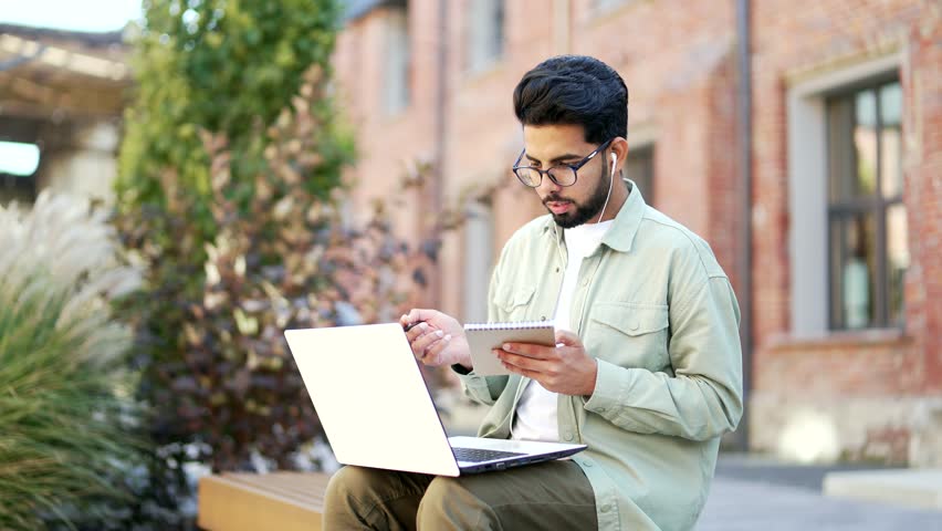 Student wearing headphones studying while watching online webinar, class webcast, looking at laptop, e-learning, taking notes, video call, prepares for university exams sitting on a bench on campus