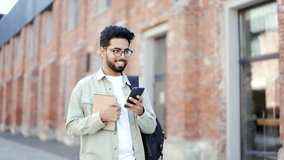 A handsome student with a backpack and books in his hands uses a smartphone walking around the campus space near university building. Smiling male chatting online, browsing social media or texting  - Powered by Shutterstock - Get 15% off with code: PIKWIZARD15