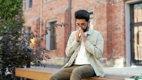 A student sneezes because he has seasonal allergies sitting on a bench in campus space near university building. Male wipes his nose with a handkerchief. He has a cold or respiratory tract infection - Powered by Shutterstock - Get 15% off with code: PIKWIZARD15