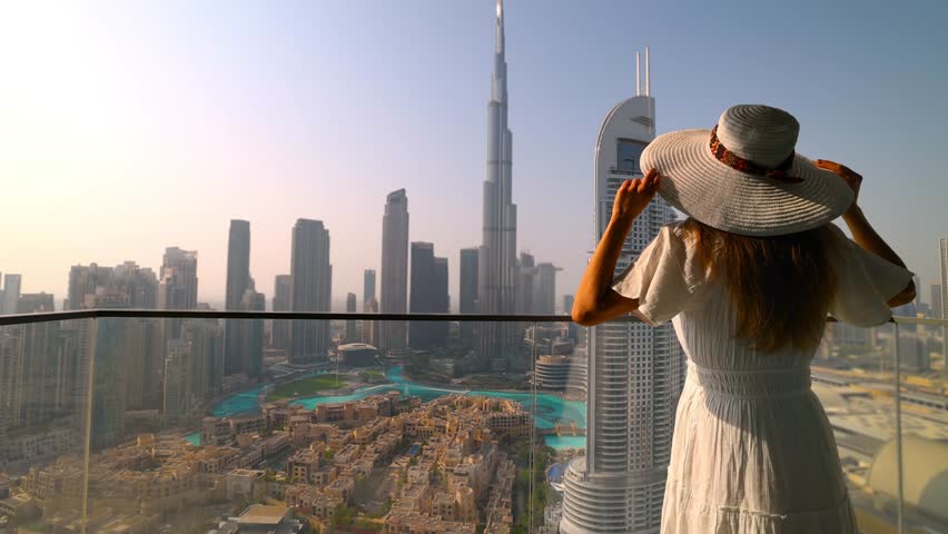 A tourist woman in a white dress looks at the sunset view of the downtown Business Bay district skyline of Dubai, UAE