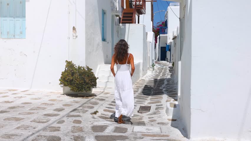 A tourist woman in a white summer dress walks through the whitewashed streets at the town of Mykonos island, Cyclades, Greece