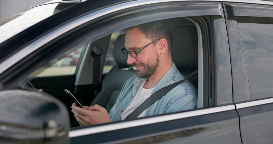 Young Caucasian man sits in a car on a parking lot and typing message on a smart phone | Shutterstock HD Video #1111813803