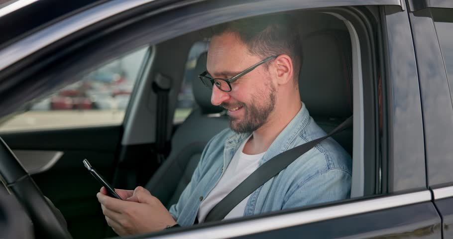 Young Caucasian man sits in a car on a parking lot and typing message on a smart phone | Shutterstock HD Video #1111813813