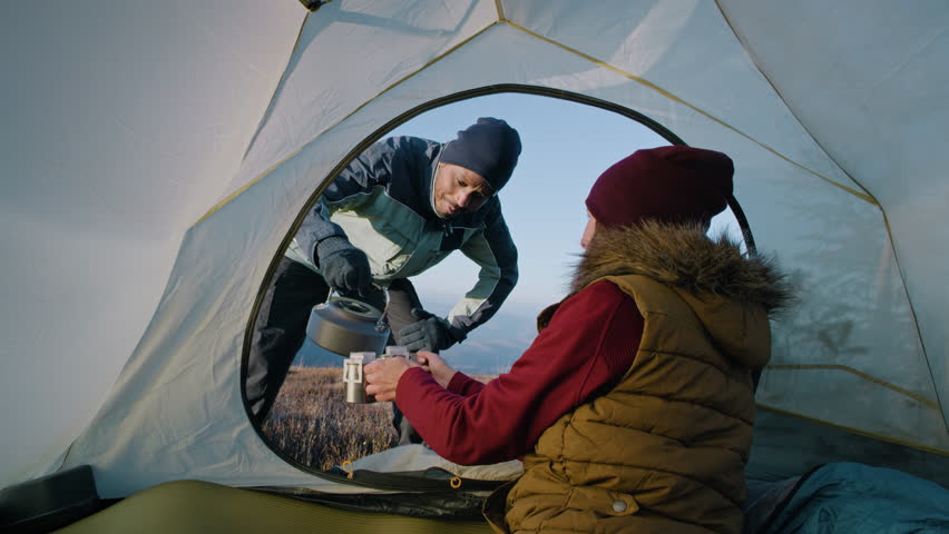 Woman sits in tent and keeps two cups with tea in hands. African American man with boiling kettle pours water in cups. Multiethnic backpacker family warm up at cold windy weather on mountain hill. | Shutterstock HD Video #1111815111