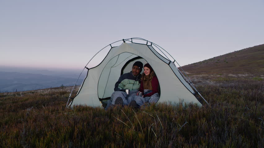 African American man with Caucasian woman sits in tent on hill and talks. Multiethnic tourists drink tea and look at sunset. Young hikers enjoy view on their vacation or trip in mountains in autumn. | Shutterstock HD Video #1111815113