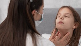 Pediatric doctor checking tonsils of a child girl. Kid health diagnostic at home. Woman doctor in face mask examining small girl patient throat. Child medical care.