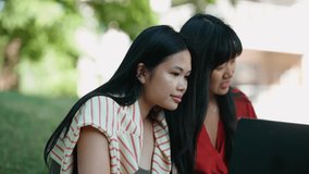 Two cute asian female students talking by video call on laptop and showing book outdoors