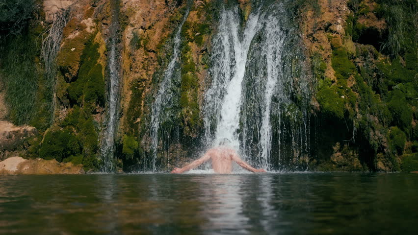 Contented man relax and swim under waterfall flowing water in mountain pond. People enjoy of nature in vacation. 
