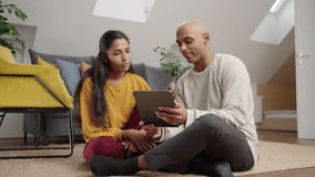 Adult couple sitting on the floor in their apartment, searching something on the tablet and having a conversation