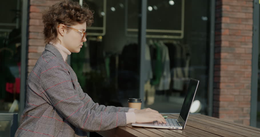 Side view of confident young woman using laptop typing and drinking coffee working in lunch break in street cafe. Business and freelance job concept. | Shutterstock HD Video #1111822035