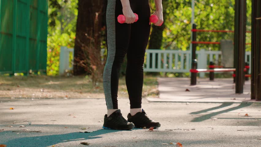 Close up view of woman wearing sport clothes and does squats with dumbbells in city park. Slow motion. Concept of healthy lifestyle and fitness