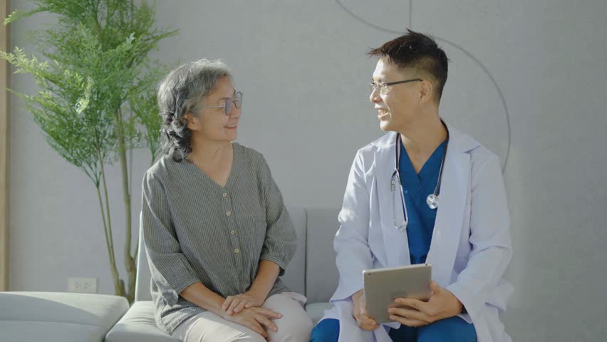 Asian young man doctor holding digital tablet consulting old patient, Male doctor therapist in uniform sit on sofa at hospital or clinic with senior patient during medical visit doing health checkup | Shutterstock HD Video #1111826689
