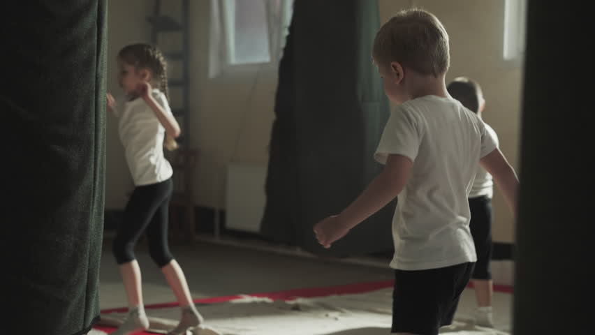 Child attacks punching bag with classmates in gym. Toddler boy beats hanging equipment by legs and hands in classroom slow motion. Kickboxing team | Shutterstock HD Video #1111829359