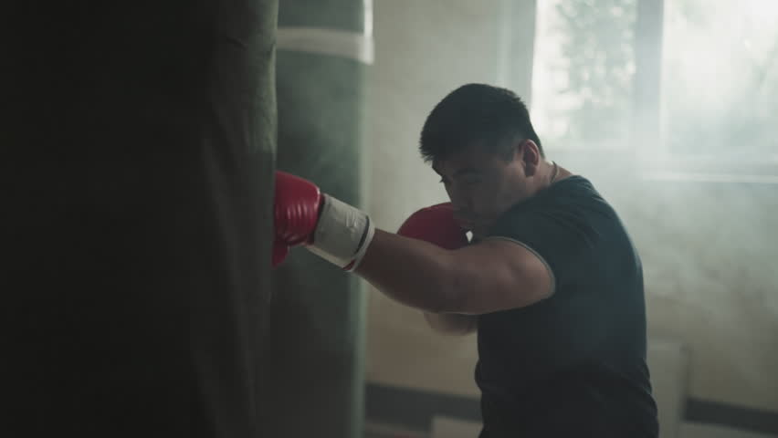 Man punches heavy bag with hooks in sports club. Professional sportsman has active training in equipped gym with dust cloud slow motion. Martial arts exercises | Shutterstock HD Video #1111829393
