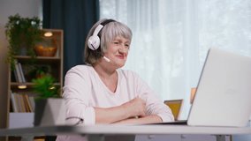 smiling woman with a headset talking on a video call using a web camera on a laptop while sitting at a table in a cozy room