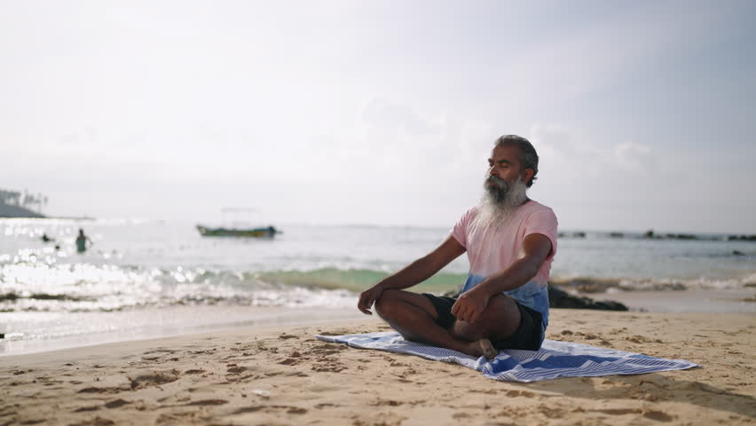 Senior grey bearded traveler use meditation practice and yoga to clear mind, sitting in lotus pose on tropical island. Elderly man with closed eyes enjoy retirement on beach with palm trees, ocean. | Shutterstock HD Video #1111830247