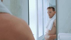 Young contented Caucasian man in towel checking his body shape in mirror in bathroom. Skinny satisfied male checking his stomach and body fat percentage in the morning - Powered by Shutterstock - Get 15% off with code: PIKWIZARD15