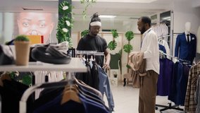 Man looking at elegant shirts in clothing store, trying to determine if they fit, asking employee for feedback. Client receiving fashion boutique garments recommendations from friendly staff member