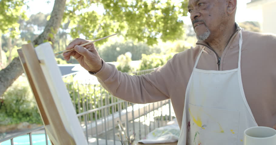 Happy senior biracial man painting at balcony at home, slow motion. Domestic life, retirement, art, nature and lifestyle, unaltered.