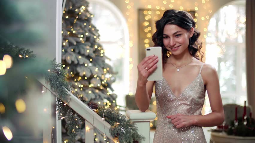 Pretty woman has a video call with beautiful Christmas decoration in the background. Brunette-haired girl smiles and talks via smartphone. High quality 4k footage
