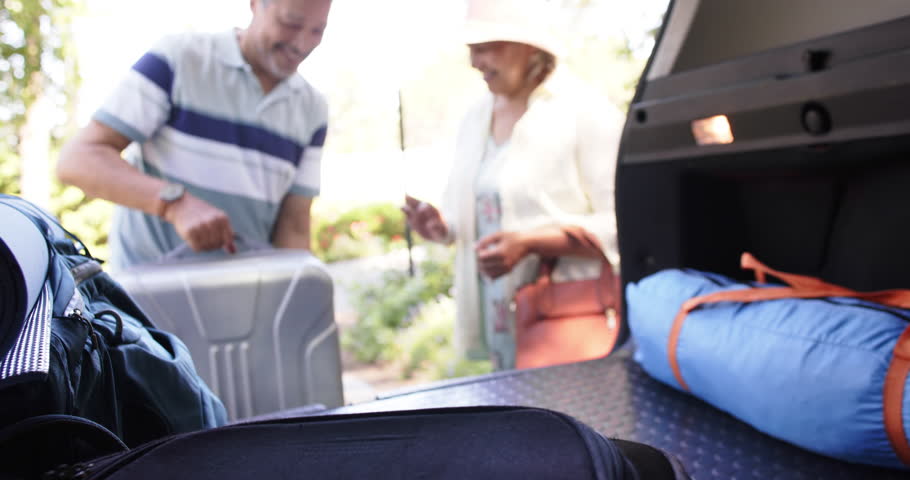 Happy diverse senior couple packing luggage to a car in sunny outdoors. Retirement, leisure, traveling and nature concept, unaltered.