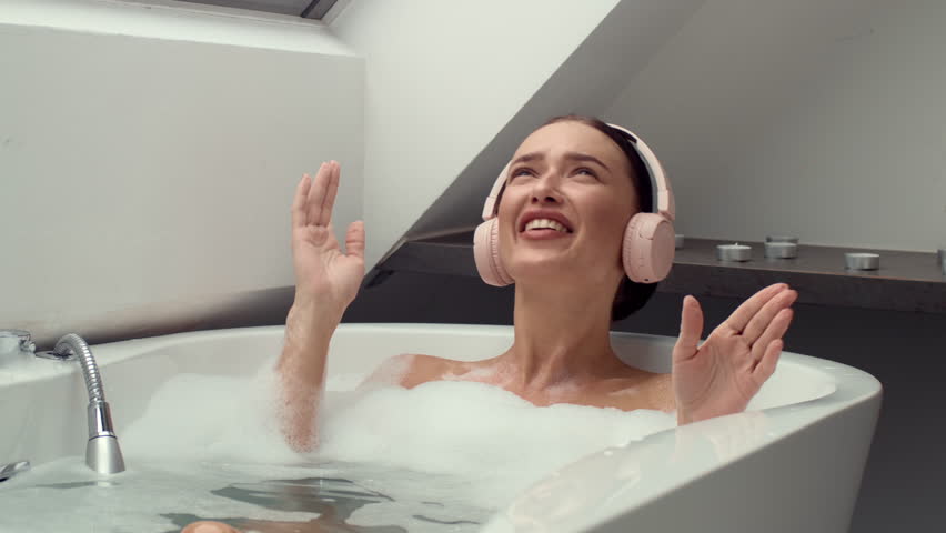 4K slow-motion footage captures a happy woman in a foamy bath, donning headphones, lost in music and singing aloud. The scene radiates relaxation, joy, and the therapeutic power of music Royalty-Free Stock Footage #1111848435