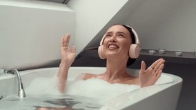 4K slow-motion footage captures a happy woman in a foamy bath, donning headphones, lost in music and singing aloud. The scene radiates relaxation, joy, and the therapeutic power of music