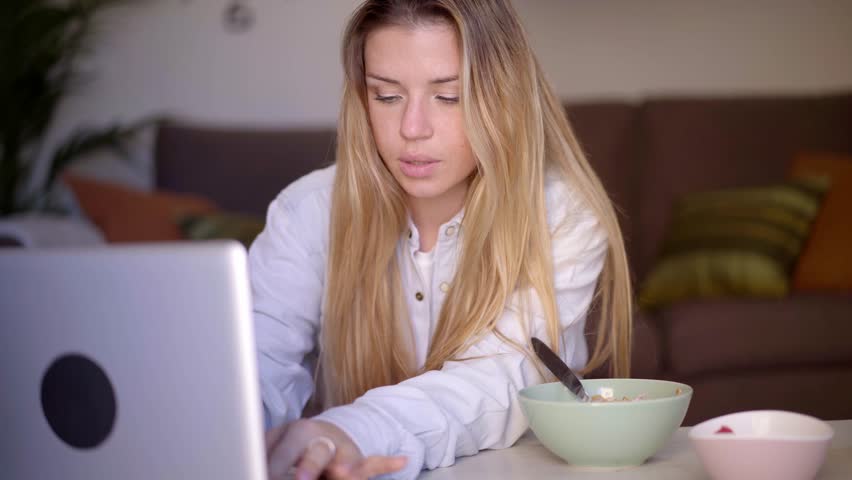 Young adult Caucasian woman eating breakfast sitting in living room concentrating on laptop screen. Blonde gen z female snacks at home on bowl of cereal with yogurt and fruit while using computer | Shutterstock HD Video #1111850561