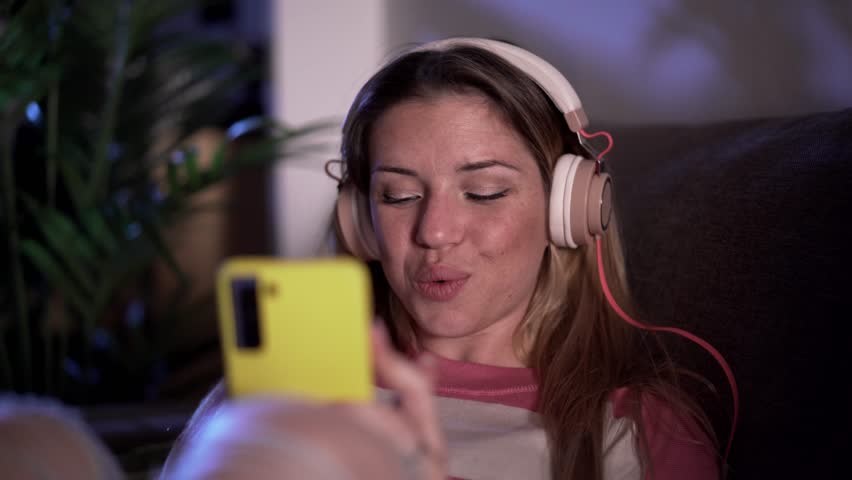 Cheerful young blonde woman listening to music with headphones sitting on sofa at home in evening. Caucasian girl using mobile phone enjoys dancing favorite song in living room. Electronic devices | Shutterstock HD Video #1111850567