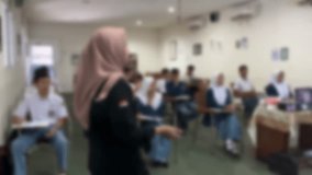 A teacher is giving a lesson. The presenter is speaking in front of the audience. Blurred classroom background.
