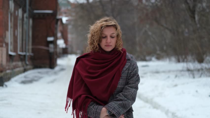 A young woman with curly hair is wearing a winter coat and a burgundy scarf. Woman on the street on a winter day. There's snow all around her | Shutterstock HD Video #1111856211