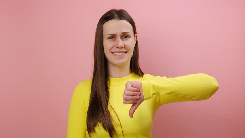 Dislike concept. Upset dissatisfied young woman showing thumb down and frowning face in disgust, negative feedback, wearing yellow sweater, posing isolated on pink color background wall in studio | Shutterstock HD Video #1111863265
