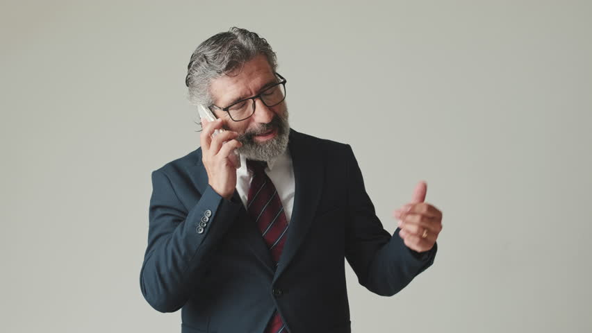 Businessman in glasses with gray hair, talking on mobile phone with smile, isolated on gray background in the studio | Shutterstock HD Video #1111864641