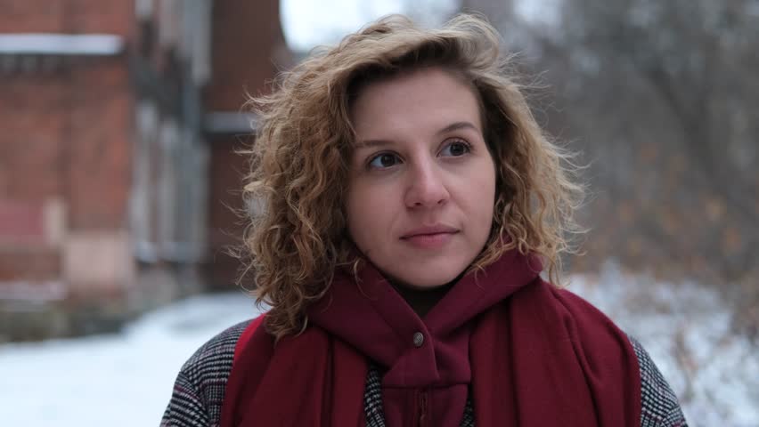 A young woman with curly hair is wearing a winter coat and a burgundy scarf. Woman on the street on a winter day. There's snow all around her | Shutterstock HD Video #1111866513