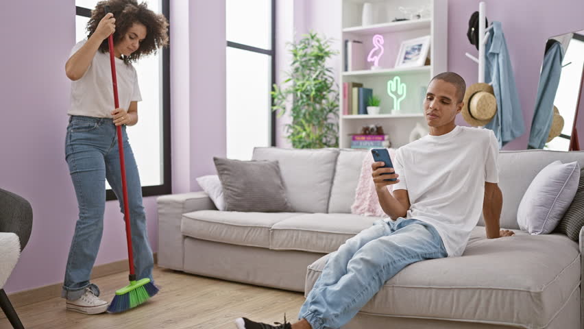 Charming couple enjoying household chores, beautiful woman cleaning floor, lovely boyfriend smiling  texting love message on smartphone at their cozy apartment indoors | Shutterstock HD Video #1111866915