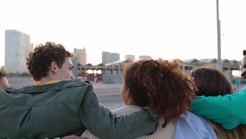 Rear view of young united group of multiracial friends walking together while hugging each other outside. Unity, youth community and international friendship concept.  Royalty-Free Stock Footage #1111867341