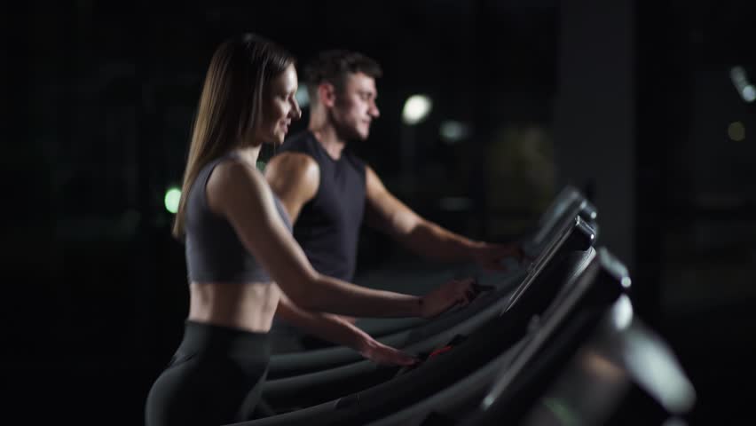 Wellness, young woman and man athlete walks and starts the run on a treadmill, two runners perform aerobic exercise and endurance training in the gym, nightlife. | Shutterstock HD Video #1111867555