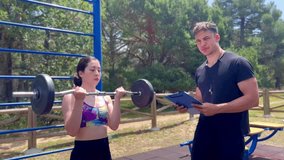 Video of portrait of a Latina woman performing biceps curls with some weights in an outdoor sports park, while her personal trainer takes notes on her chart and monitors her training