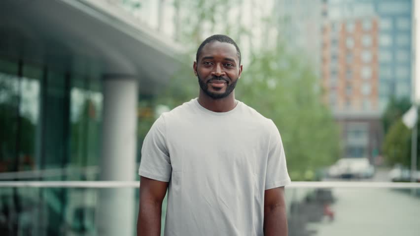 Portrait smiling positive african american bearded man standing on ctiy street in closed pose with hands on chest looking at camera. Friendly guy in t-shirt in downtown. Human emotions, body language. | Shutterstock HD Video #1111871637
