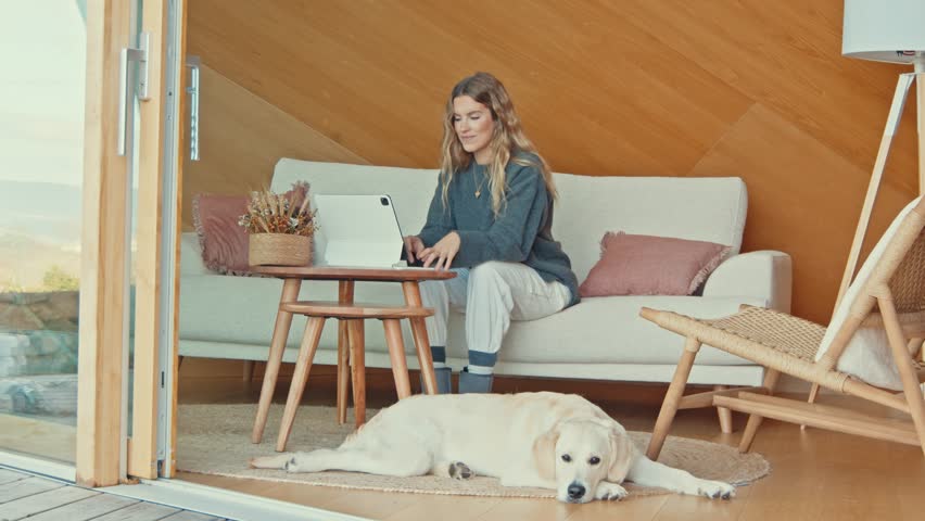 Cheerful woman using a tablet with her dog by her side in a wooden home. | Shutterstock HD Video #1111872401
