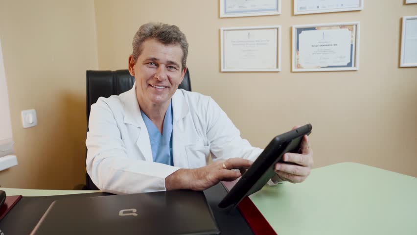 A doctor uses a tablet while sitting at a desk in his office. | Shutterstock HD Video #1111873491